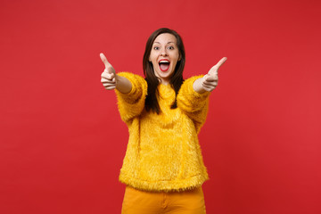 Excited young woman in yellow fur sweater keeping mouth wide open, showing thumbs up isolated on bright red wall background in studio. People sincere emotions, lifestyle concept. Mock up copy space.