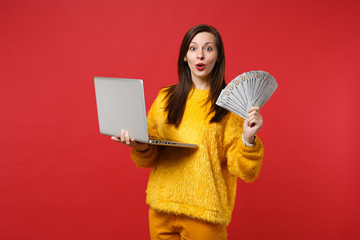 Amazed young woman in yellow fur sweater holding laptop pc computer, fan of money in dollar banknotes, cash money isolated on red background. People emotions, lifestyle concept. Mock up copy space.