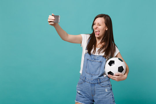 Funny young girl football fan with soccer ball doing selfie shot on mobile phone showing tongue isolated on blue turquoise background. People emotions sport family leisure concept. Mock up copy space.