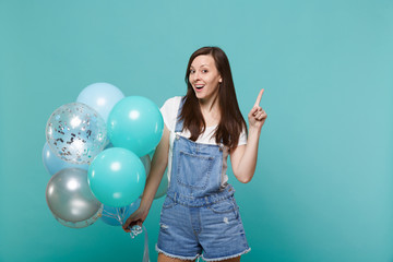 Amazed young woman in denim clothes pointing index finger up, celebrating and holding colorful air balloons isolated on blue turquoise wall background. Birthday holiday party, people emotions concept.