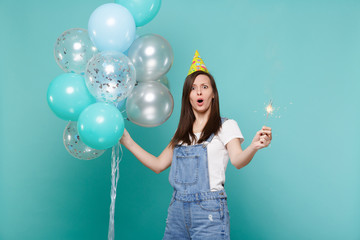 Perplexed woman in birthday hat keeping mouth open, hold burning sparkler celebrating with colorful air balloons isolated on blue turquoise background. Birthday holiday party, people emotions concept.