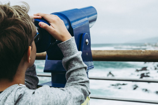 Close up view of a child looking horizon through blue telescope spyglass in front of ocean what we see far away watching the moon stars watch birds planes sky young boy looks through binoculars sea 
