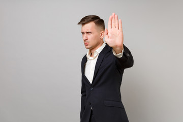Concerned young business man in classic black suit and shirt showing stop gesture with palm isolated on grey wall background in studio. Achievement career wealth business concept. Mock up copy space.