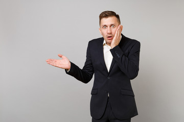 Shocked young business man in classic black suit white shirt pointing hand aside putting hand on cheek isolated on grey wall background. Achievement career wealth business concept. Mock up copy space.