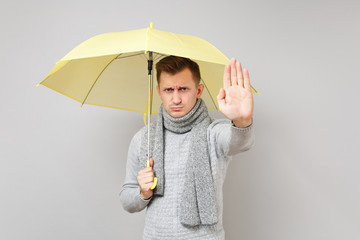Dissatisfied young man in gray sweater, scarf holding yellow umbrella showing stop gesture with...