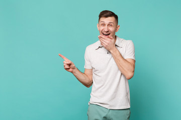Laughing young man in casual clothes pointing index finger aside and covering mouth with palm isolated on blue turquoise wall background. People sincere emotions lifestyle concept. Mock up copy space. © ViDi Studio