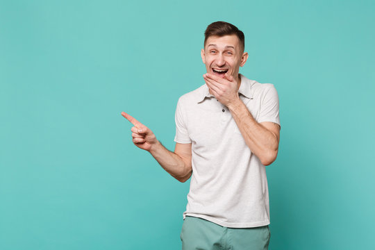 Laughing young man in casual clothes pointing index finger aside and covering mouth with palm isolated on blue turquoise wall background. People sincere emotions lifestyle concept. Mock up copy space.