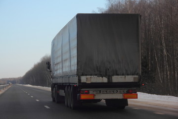 Logistics, international freight by road - dirty white european truck with semi trailer drive next to the two-lane asphalted suburban highway in the winter day, side rear view