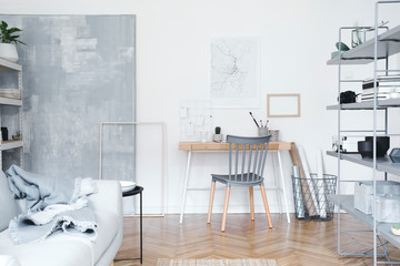 Stylish nordic home interior with creative wooden desk, grey bookstand with accessories, design grey sofa and modern furniture. Open space and living room for artist, designer. Mockup poster frame.