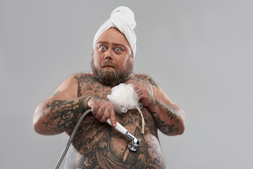 Funny tattooed man putting shower head on the belly and looking surprised