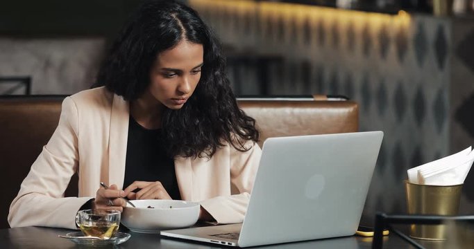 Business woman sitting with a laptop in the cafe and eating dinner with a cup of tea on the table. Eating lunch at work desk