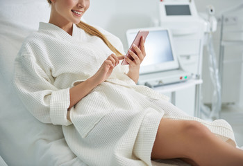 Young lady using cellphone and smiling while resting in spa salon