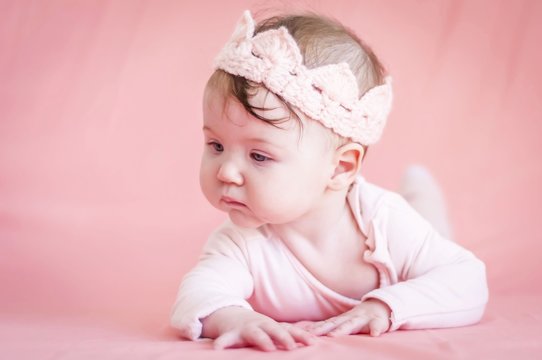 Sweet adorable six months old Caucasian baby girl with a pink cloth crown on a pink background. Infant girl revealing the world and learning to turn and crawl concept image.