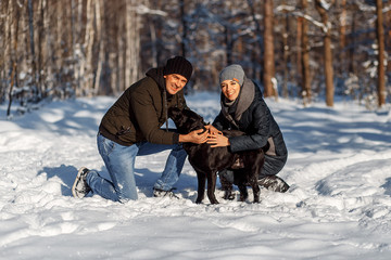 A happy couple with a dog (black Labrador) having fun outside in the forest on a sunny frosty winer...