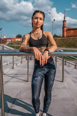 Girl athlete, summer city. Posing against the background of steps. In leggings sneakers. Woman with tattoos. Confident look. Emotions of a strong and confident person.