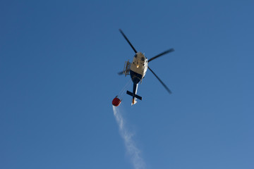 Helicóptero de extinción de fuegos de montes, dirigiendose a un fuego derramando agua de una canasta que transporta.