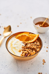 Bowl of homemade granola with yogurt, honey and fresh orange on white abstract background. Healthy breakfast. Golden utensils. Top view. Copy space for text.