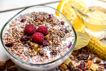 transparent glass plate with muesli sprinkled with seeds and fresh raspberries stands on the table next to a mug of green tea, honeycomb and lemon slices