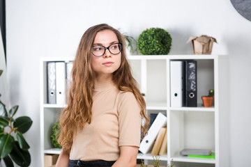 Portrait of a young Caucasian woman with long hair and glasses for friction stands in an office...