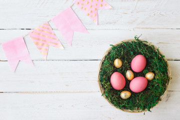 Easter. Pink and gold Easter eggs lie in a nest with moss on a white wooden background with a decoration of paper garland. Spring, April. Selective focus