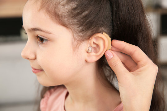 Young woman putting hearing aid in daughter's ear indoors, closeup