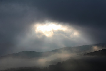 Sunbeams push through storm clouds, lighting up the mountain forest of Carpathians in fog..