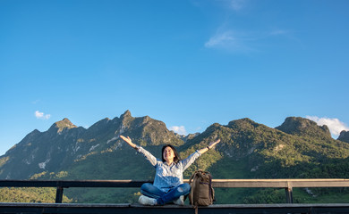 Asian woman traveler  sitting and arms up in the air at view point terrace at landscape view of mountain with cloud and blue sky in sunny day at forest.backpacker explore nature life.