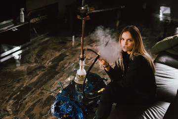 Fototapeta na wymiar Young caucasian blonde girl smoking hookah at luxury interior with custom v8 car engine table. Leather sofa and beautiful smile