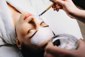Lovely woman with closed eyes lying on spa bed while on face is applying white mask with a brush in a spa salon.