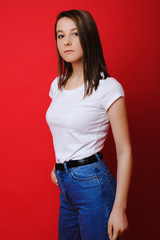 Portrait of adorable brunette posing seriously at the camera dressed in white shirt and blue jeans against a red wall.