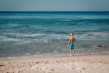 young boy  in blue shorts and blue sunglass stands at shore and watches waves in ocean, back view looking out. Phuket. Thailand.