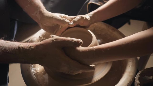 Two intertwined hands form a pot of clay on a potter's wheel. A male hand helps a woman's hands shape a clay shape.