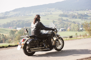 Long-haired bearded biker in sunglasses and black leather outfit driving cruiser powerful motorcycle along sunny asphalt road on bright summer day on background of green rural misty landscape.