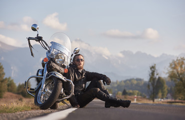 Handsome motorcyclist in black leather clothing sitting at modern powerful shiny motorcycle on grassy roadside on blurred copy space background of distant mountain peaks under bright sky.