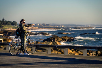 Young and beautiful girl is riding a bike by pier next to the Atlantic ocean during sunset time. Beautiful light. Smiley face. Porto, Portugal. - 247119500