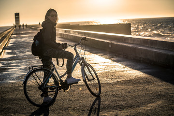 Young and beautiful girl is riding a bike by pier next to the Atlantic ocean during sunset time. Beautiful light. Smiley face. Porto, Portugal. - 247119559