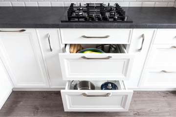 Opened drawers on a white black classic traditional kitchen with kitchen utensils inside. 