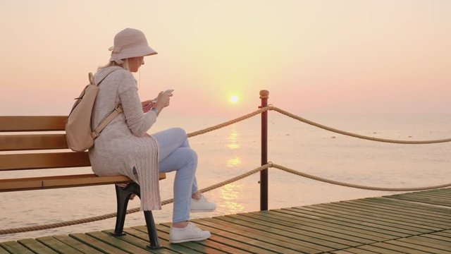 A stylish girl in a pink hat sits on a bench on a sea pier and reads a text message on the phone, the rising sun is reflected in the blue water, coloring the sky in pink