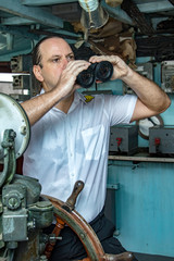 The helmsman stands in the wheelhouse and watching the surroundings. A sailor officer in white uniform looking to sea with binocular. © milkovasa