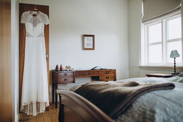 white wedding dress hanging on a wooden closet in a bright bedroom with a large white window, next to the bride's shoes are on the table