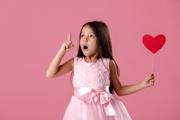 portrait of cute little girl in pink dress holding paper heart, pointing up and having idea on pink...