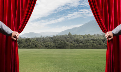 Green meadow behind red curtain and hand holding it
