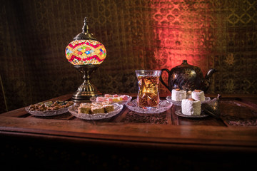 Arabian tea in glass with eastern snacks on vintage wooden surface. Eastern tea concept. Low light lounge interior with carpet. Empty space.