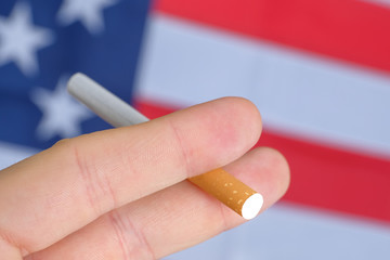 Fingers holding a cigarette on the background of the American flag. Concept of nicotine addiction problems in the US.