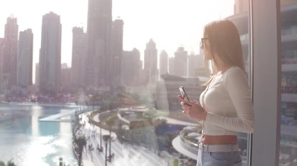 Young woman typing on the phone at the big window and looking on the Dubai city buildings. Sunset.