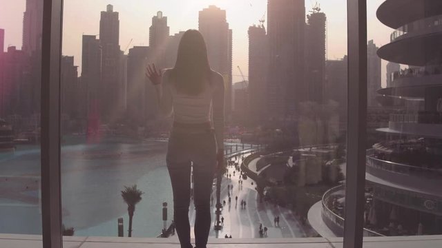 Young woman coming to the window and looking and looking down to the lake and skyscrapers at the subset.