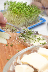 Męska dłoń układa obcięte kiełki rzeżuchy za pomocą widelca na kromce chleba posmarowanej masłem.