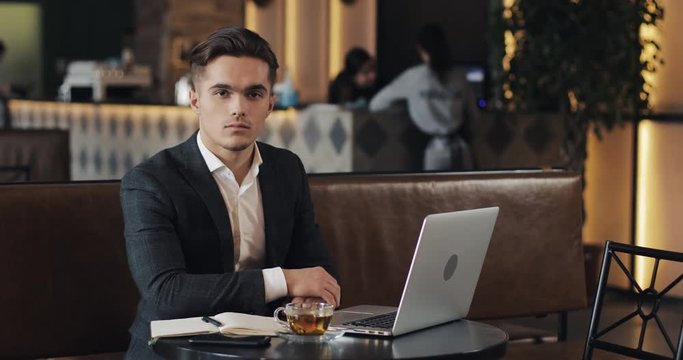 Portrait of young successful businessman sitting in cafe with laptop and looking into the camera