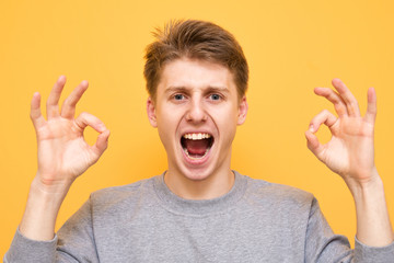 Close portrait of a boy on a yellow background shows an OK sign. Emotional young man shows an OK sign, isolated on a yellow background.