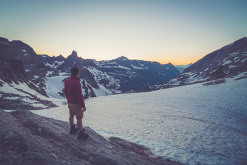 Fototapeta premium Young man standing on top of cliff in summer mountains at sunset and enjoying view of nature